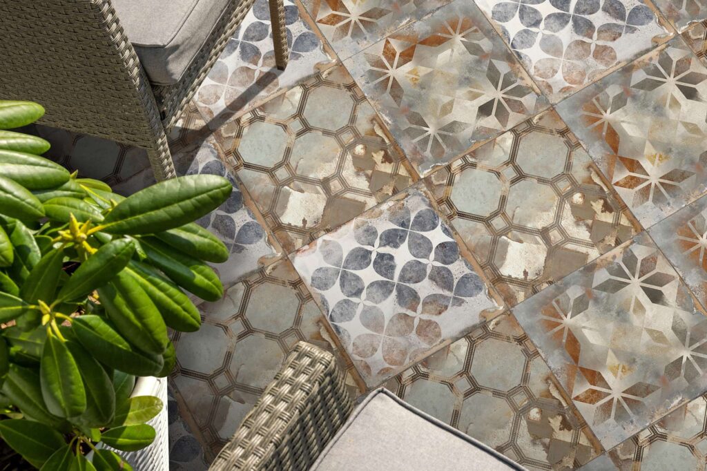 La collezione Heartland nel formato Decori 30x30 valorizza questo spazio esterno con un pavimento in gres porcellanato dall'estetica vintage e vissuta. Il close-up mostra la posa di piastrelle decorate con vari motivi geometrici e floreali, caratterizzati da sfumature di azzurro, ruggine, grigio e beige. L'atmosfera raffinata dell'outdoor è arricchita da sedie in rattan grigio con cuscini coordinati e da una pianta verde rigogliosa lambita dalla luce del sole.