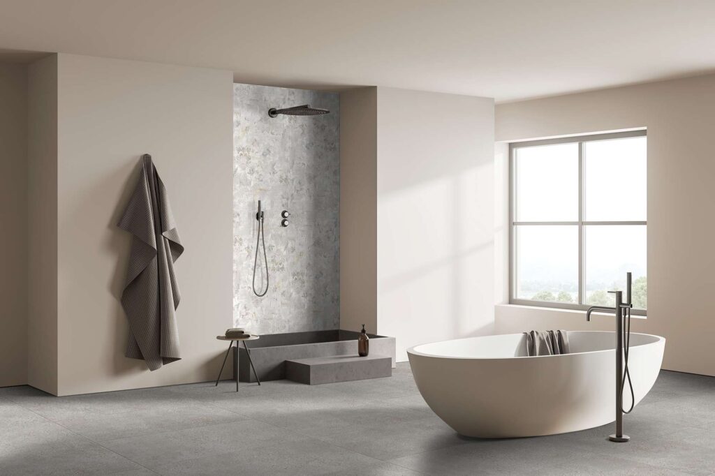 La collezione I, con il decoro Boheme nel formato 60x120, impreziosisce la parete doccia di un bagno moderno dal design raffinato e minimale. La scena mostra una spaziosa sala da bagno caratterizzata da un'atmosfera serena e luminosa, con un'elegante vasca freestanding bianca in primo piano e pavimenti in gres grigio scuro. Sullo sfondo, all'interno della nicchia doccia, risalta il rivestimento ceramico decorato Boheme, che con il suo delicato motivo floreale d'ispirazione artistica crea un sofisticato punto focale in perfetta armonia con la luce naturale che entra dalla grande finestra.