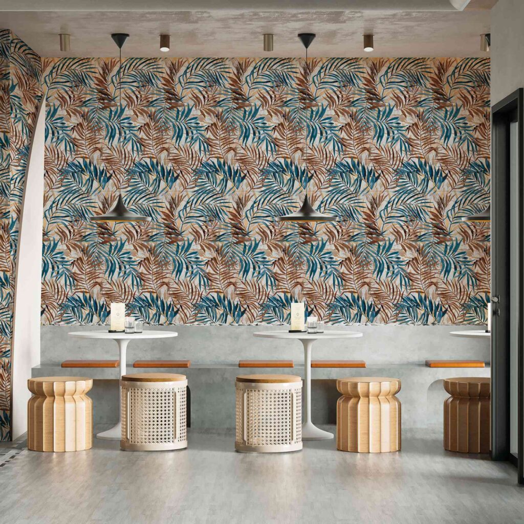 La collezione I, con il decoro Savage Steel nel formato 60x120 cm, valorizza la parete di un elegante bar moderno con un suggestivo rivestimento ceramico dall'effetto carta da parati. Le grandi lastre presentano un fitto pattern botanico con foglie tropicali nei toni del blu acciaio, del marrone bruciato e del beige, creando un forte impatto visivo e naturale. L'ambiente, caratterizzato da un'atmosfera raffinata ed esotica, è completato da tavoli rotondi bianchi, sgabelli di design in legno e rattan, una panca in cemento e un pavimento grigio chiaro che esalta la vivacità della parete decorata.
