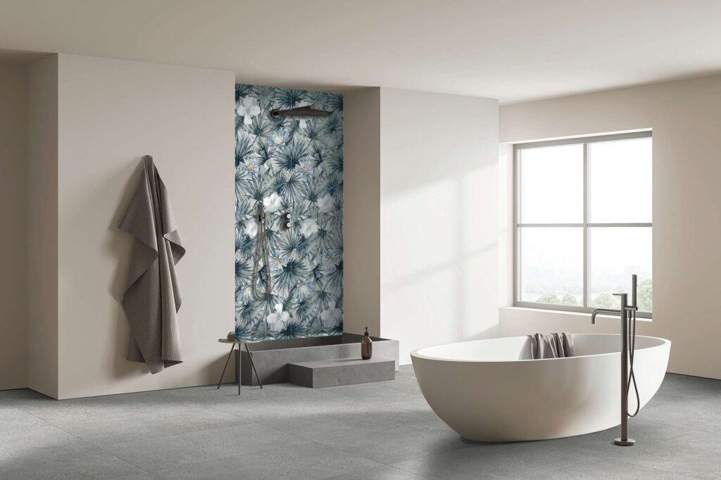 La collezione I veste un elegante e moderno ambiente bagno, dove spicca il contrasto armonioso tra natura e minimalismo. All'interno della doccia, la parete di fondo è rivestita con il decoro floreale Decors Maui Sea nel formato 60x120 cm, caratterizzato da un raffinato motivo di foglie tropicali e fiori bianchi su fondo blu-grigio. A pavimento, la posa delle piastrelle Mon Smoke nel formato 60x120 cm offre una superficie materica grigia che dona sobrietà e continuità visiva all'ambiente. Lo spazio, ampio e inondato di luce naturale grazie a una grande finestra, ospita una vasca freestanding bianca di design, evocando un'atmosfera di puro benessere e raffinata spa domestica.