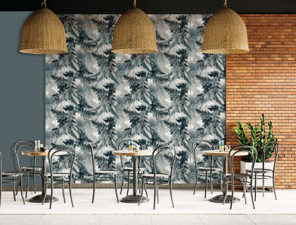 La collezione I presenta il decoro Proton Texdeco Cold nel formato 60x120, posato come suggestivo rivestimento a parete in un ristorante dall'atmosfera moderna e accogliente. Le piastrelle ceramiche creano una carta da parati ceramica dal raffinato motivo botanico con grandi foglie tropicali nei toni freddi del grigio, blu polvere e verde salvia. La scena mostra un'elegante sala da pranzo con tavoli rotondi in legno, sedie metalliche dallo stile rétro, grandi lampadari a sospensione in paglia intrecciata e una parete laterale con mattoni a vista che riscalda l'ambiente.