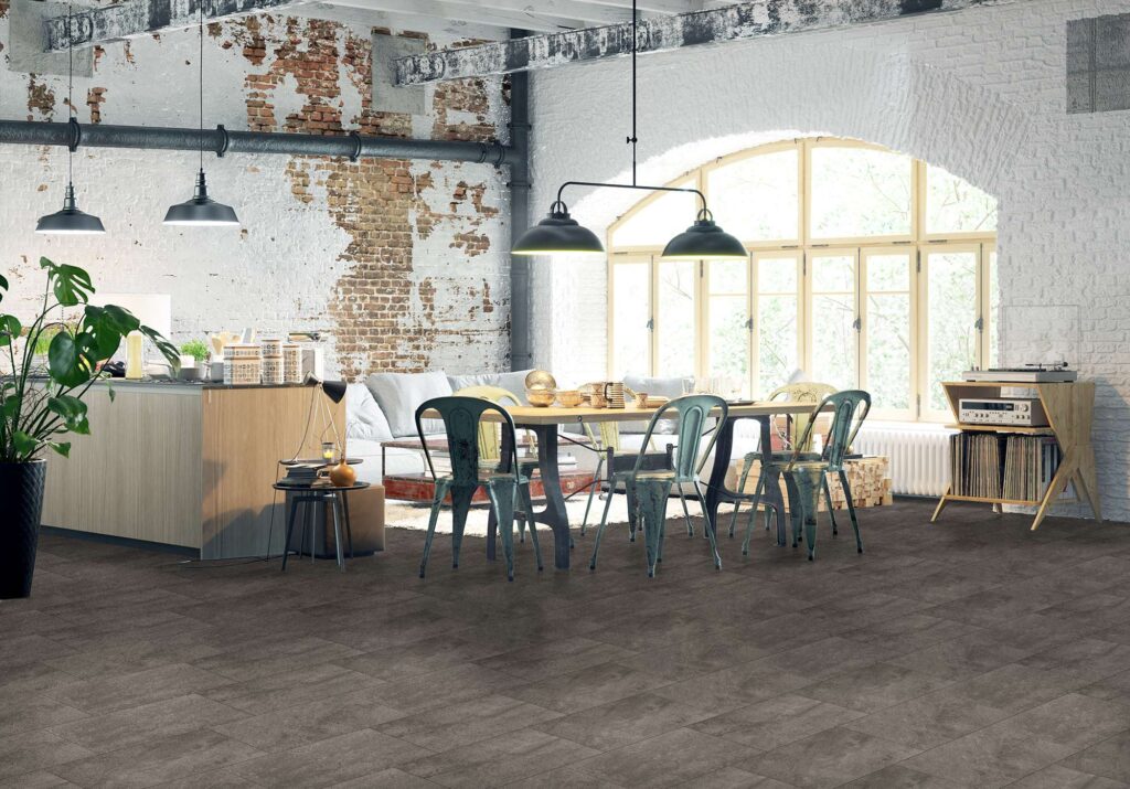 La collezione di piastrelle Jelling Grey nel formato 30x60 cm riveste il pavimento di un ampio e luminoso living in stile loft industriale. Le piastrelle in gres porcellanato grigio scuro, caratterizzate da una texture materica e posate a correre, si integrano elegantemente in un ambiente caratterizzato da una parete con mattoni a vista parzialmente intonacati, un tavolo da pranzo in legno con sedie metalliche dal design vintage e una grande finestra ad arco che inonda lo spazio di luce naturale.
