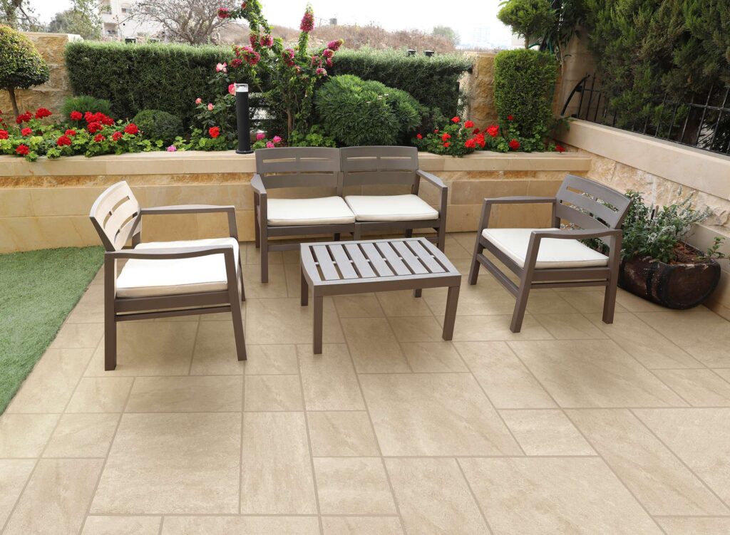 La collezione Jelling Sand, nella calda tonalità sabbia, valorizza questo elegante spazio outdoor con un pavimento in gres porcellanato effetto pietra naturale. Le piastrelle, posate in uno schema modulare multiformato che combina le dimensioni 60x60, 30x60 e 30x30 cm, creano una superficie dinamica e accogliente. Sul patio è disposto un raffinato salottino da giardino composto da divano, poltrone e tavolino marroni con cuscini color crema, circondato da una rigogliosa siepe verde impreziosita da piante fiorite e rose rosse.