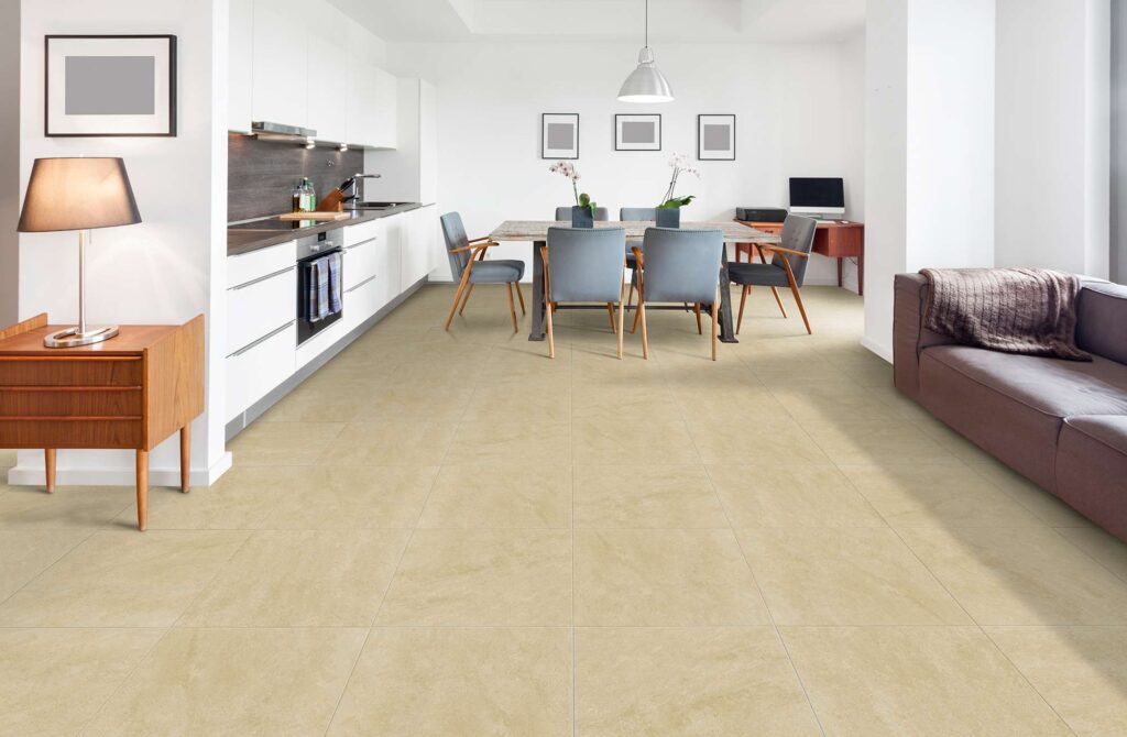 La collezione Jelling Sand, nel formato 60x60 cm, veste il pavimento di un luminoso e moderno open space. Le piastrelle in gres porcellanato nei toni caldi del sabbia, caratterizzate da una texture naturale e posate in modo regolare, collegano armoniosamente la cucina lineare bianca sulla sinistra, l'elegante zona pranzo centrale con sedie grigie e l'area living sulla destra con divano, definendo un'atmosfera d'insieme accogliente e dal design contemporaneo.
