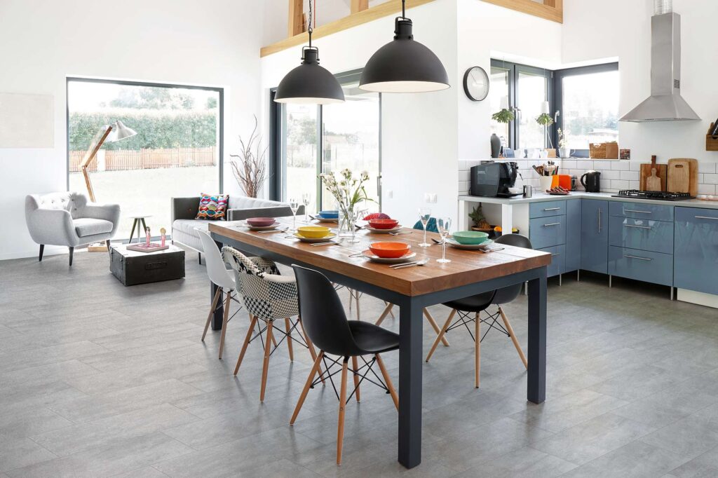 Pavimento in gres porcellanato della collezione Jelling Silver nel formato 30x60 cm, caratterizzato da eleganti piastrelle grigie effetto pietra con posa a correre. La superficie valorizza una spaziosa e luminosa cucina open space con zona pranzo, arredata con un grande tavolo in legno massiccio, sedie di design contemporaneo e dettagli moderni, che si apre su un'accogliente area living con ampie vetrate sul giardino.
