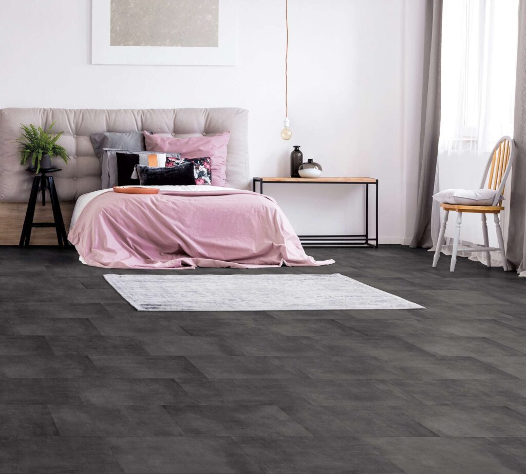 La collezione Materia in gres porcellanato color Antracite, nel formato 30x60, riveste il pavimento di un'elegante e luminosa camera da letto. La posa sfalsata mette in risalto la superficie scura e dall'aspetto materico, creando un raffinato contrasto con gli arredi dai toni soft: un letto matrimoniale con testiera imbottita beige e morbidi tessili rosa cipria, un tappeto chiaro, una consolle in legno e una sedia accostata alla finestra da cui filtra una calda luce naturale.