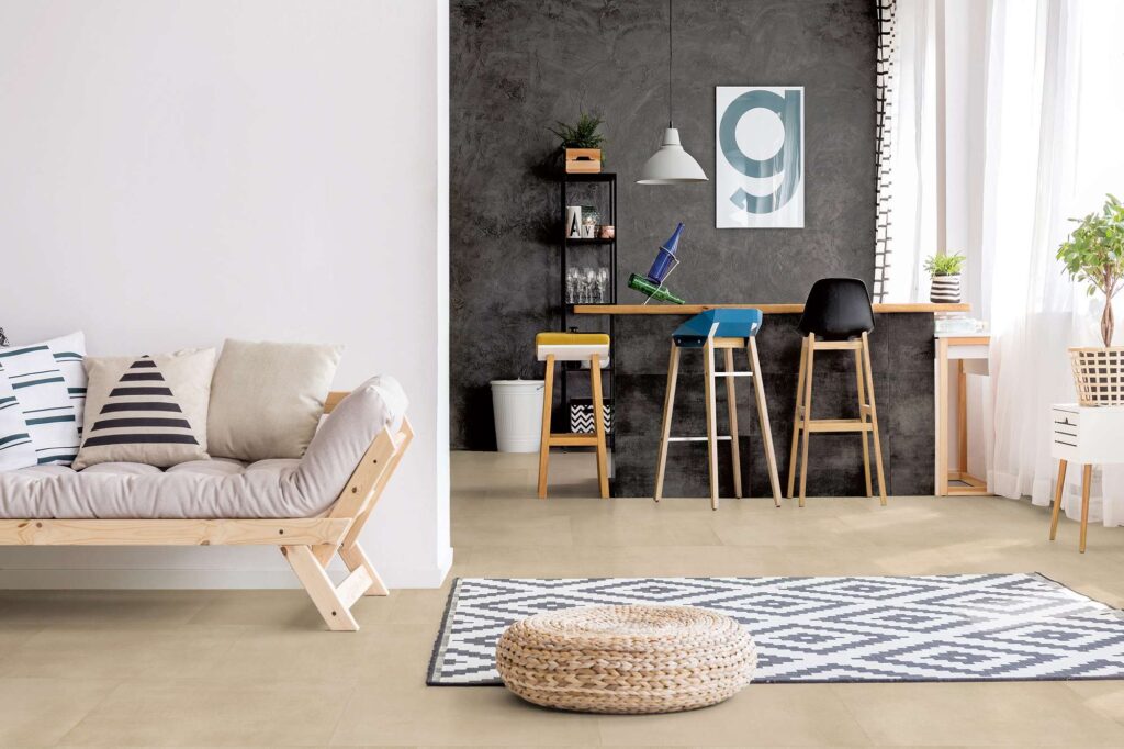 La collezione Materia nella tonalità Beige e formato 60x60 cm riveste il pavimento di un moderno e luminoso open space di design. Le piastrelle in gres porcellanato, caratterizzate da una tonalità calda e naturale, sono posate in modo regolare e continuo in tutta la stanza. L'ambiente unisce un'area relax in primo piano, completa di divano in legno chiaro, tappeto con pattern geometrico e pouf in paglia intrecciata, a una zona bar sullo sfondo, valorizzata da una parete scura a contrasto ed eleganti sgabelli alti.