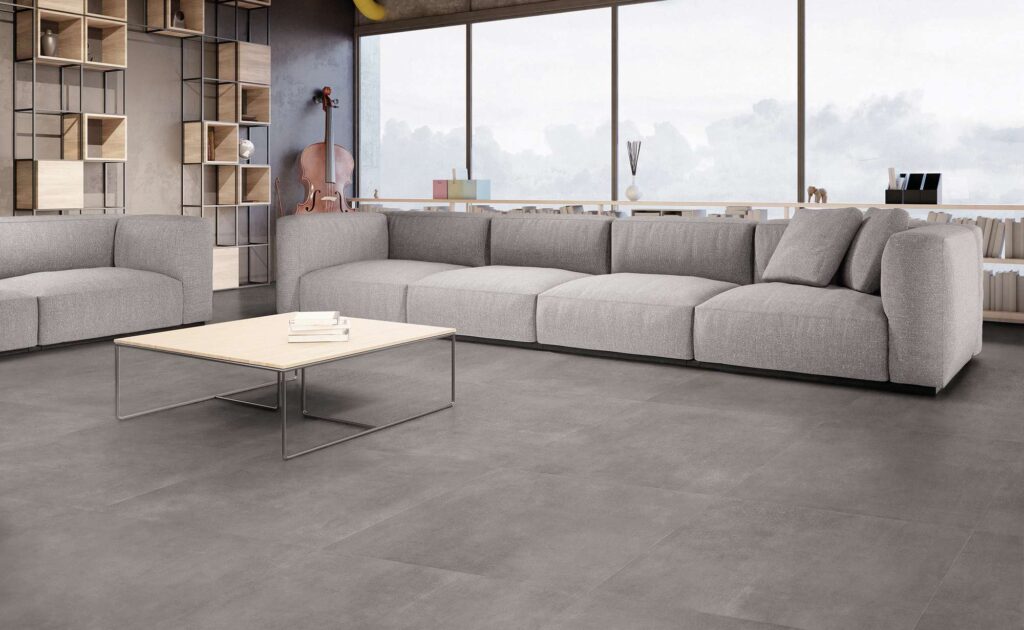 La collezione Materia nella tonalità Grafite, nel formato 60x60 cm, riveste il pavimento di un raffinato e luminoso ambiente living dal design contemporaneo. Le piastrelle in gres porcellanato grigio scuro, caratterizzate da una texture materica e posate con precisione, fanno da cornice a un arredamento moderno e minimalista: un ampio divano modulare grigio chiaro, un tavolino basso con top in legno e un'elegante libreria a giorno in metallo e legno. Sullo sfondo, una grande vetrata a tutta altezza inonda lo spazio di luce naturale, valorizzando l'atmosfera sobria e sofisticata della stanza.