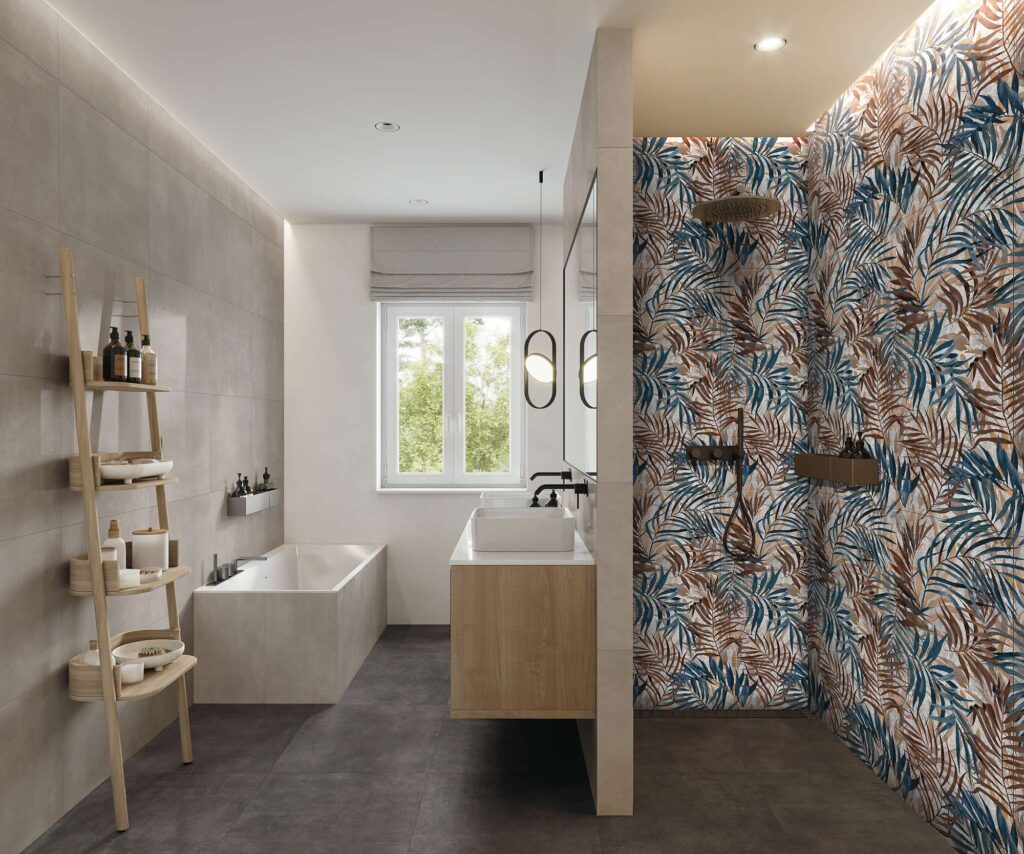 La collezione Materia nei toni Grafite e Antracite, posata a pavimento nel formato 60x60, definisce il carattere moderno e raffinato di questo bagno di design, abbinandosi al rivestimento decorativo I_Decors Savage Steel nel formato 60x120. L'ambiente, luminoso e contemporaneo, presenta un pavimento scuro in gres porcellanato, pareti grigio chiaro effetto cemento che incorniciano una vasca da bagno integrata, un mobile lavabo sospeso in legno e, sulla destra, una suggestiva doccia walk-in caratterizzata da una parete d'accento con un elegante motivo tropicale di foglie nei toni del blu, bronzo e ruggine.