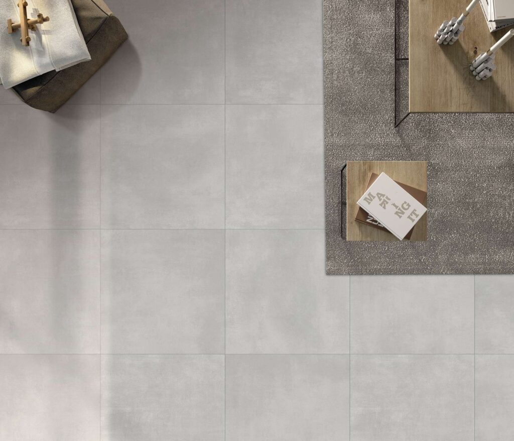 La collezione Materia, nella tonalità Grigio e formato 60x60 cm, riveste il pavimento di un moderno soggiorno in questa raffinata veduta dall'alto. Le piastrelle in gres porcellanato, caratterizzate da una texture minimalista effetto resina, sono posate in linea per un risultato armonioso e contemporaneo. Sulla destra, la superficie è parzialmente coperta da un tappeto grigio materico su cui poggiano tavolini in legno con libri di design, mentre sulla sinistra un pouf marrone con dettagli in tessuto completa un'atmosfera calda e accogliente.