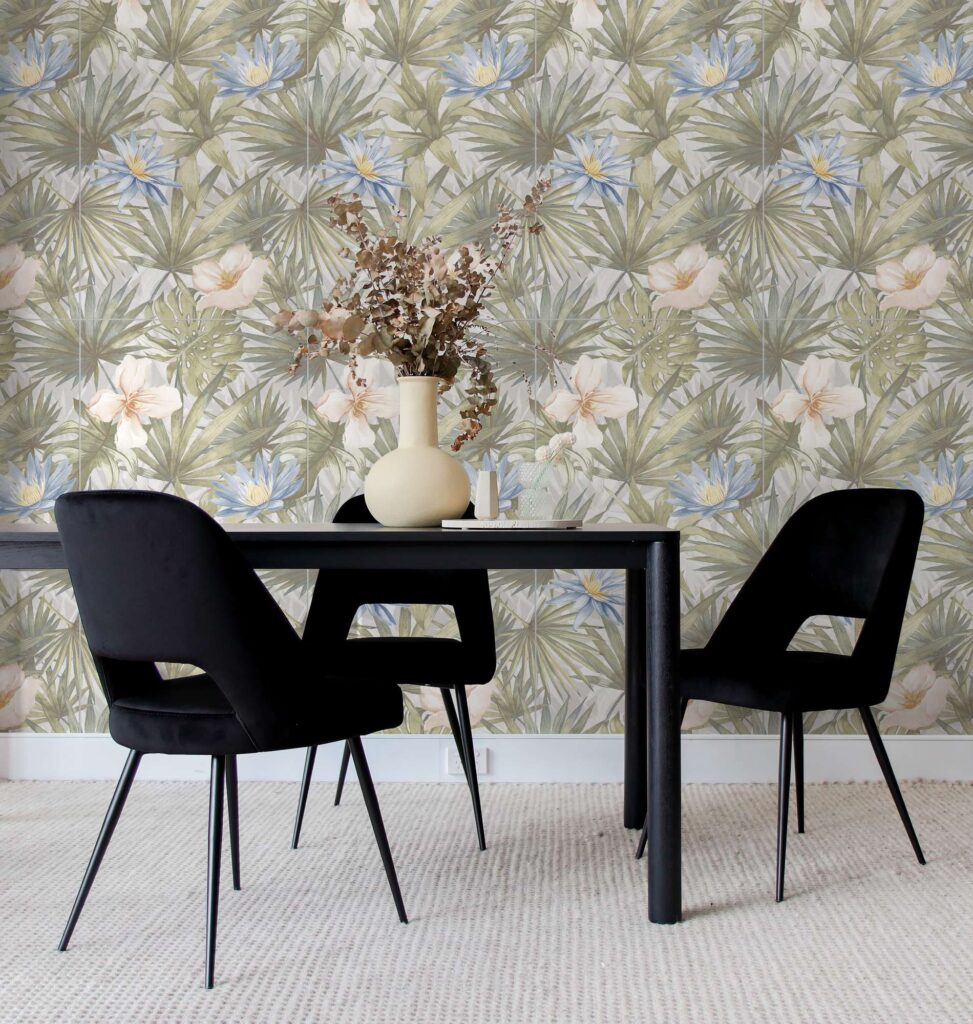 La collezione di piastrelle MAUI SUN in formato 60x120 cm valorizza la parete di questo elegante spazio living con un decoro botanico di ispirazione tropicale, caratterizzato da sfumature di verde, fiori azzurri e tonalità neutre calde. In un'atmosfera moderna e accogliente, la posa a tutta parete a effetto carta da parati fa da sfondo a una raffinata zona pranzo arredata con un tavolo scuro, sedie di design in velluto nero e un vaso in ceramica con rami decorativi.