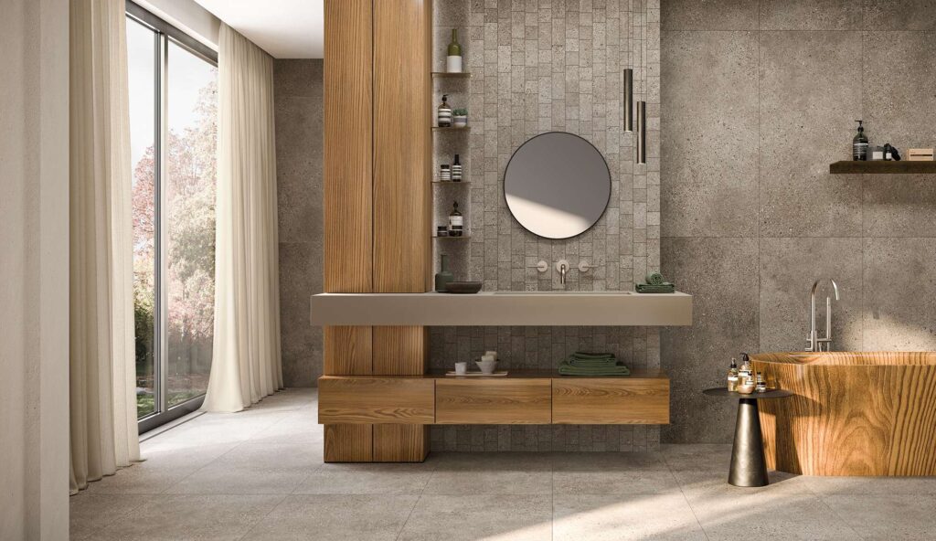 La collezione Mon Greige, proposta nei formati 60x120 e nel raffinato mosaico MOS.BRICK, definisce l'atmosfera di un bagno di lusso dal design caldo e naturale. Il gres porcellanato effetto pietra nella tonalità greige riveste con continuità il pavimento e le pareti, dove spicca la posa a mattoncino del mosaico dietro il moderno lavabo sospeso. L'arredo si distingue per i dettagli in legno naturale, come la colonna contenitrice e l'esclusiva vasca freestanding, inondati dalla luce soffusa che filtra da un'ampia vetrata con tende leggere.