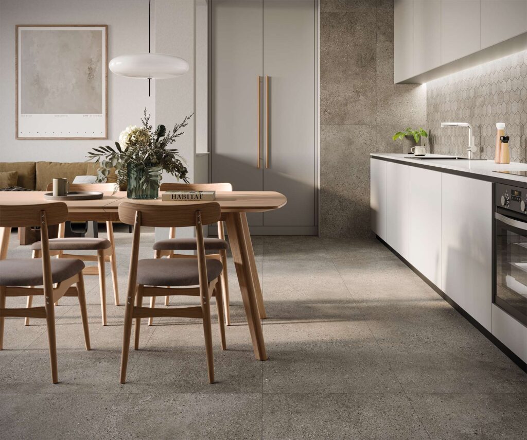 La collezione Mon Greige in gres porcellanato effetto pietra arreda una cucina moderna e luminosa, con pavimenti e rivestimenti verticali nel formato 60x120 cm nella calda tonalità greige. Sullo sfondo, la zona lavoro con mobili bianchi minimalisti è valorizzata dal paraschizzi in mosaico esagonale coordinato (Mos. Esagona). Sulla sinistra, la zona pranzo presenta un tavolo e sedie in legno chiaro, definendo un'atmosfera calda, elegante e contemporanea, esaltata dalla luce naturale che mette in risalto la raffinata matericità delle superfici ceramiche.