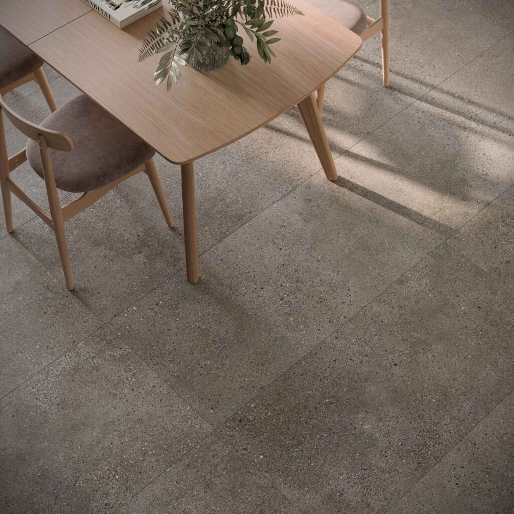 La collezione MON GREIGE nel formato 60x120 cm veste il pavimento di una raffinata zona pranzo adiacente alla cucina. Le grandi lastre in gres porcellanato effetto pietra in tonalità greige, caratterizzate da una texture materica finemente puntinata, valorizzano l'ambiente accogliendo un tavolo in legno chiaro e sedie coordinate dal design contemporaneo. La luce naturale che filtra trasversalmente esalta i dettagli della posa e la superficie elegante e naturale delle piastrelle.