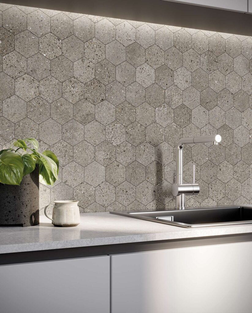 La collezione MON GREIGE MOS.ESAGONA presenta un raffinato mosaico esagonale effetto pietra nei toni caldi e naturali del greige, ideale come rivestimento per una cucina elegante e contemporanea. Nell'immagine, il backsplash in mosaico è valorizzato da una luce LED calda posizionata sotto il pensile, che mette in risalto la texture materica e i dettagli minerali delle singole tessere. Sul piano di lavoro chiaro e luminoso sono disposti un moderno lavello con miscelatore di design in acciaio inox, una tazza in ceramica artigianale e una pianta verde in vaso, ricreando un'atmosfera d'interni accogliente e sofisticata.