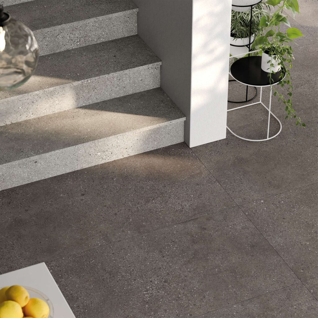 La collezione in gres porcellanato Mon Moka, nel formato 60x120, definisce lo stile di questo raffinato ambiente living. L'immagine mostra un dettaglio della pavimentazione effetto pietra nella tonalità scura Moka, caratterizzata da una texture materica e naturale, accostata a una scala interna rivestita con gradini grigio chiaro nella variante Smoke. La luce del sole taglia trasversalmente la scena, esaltando i dettagli minerali delle superfici. Sulla destra, un elegante portavaso di design con piante verdi rampicanti e un accenno di parete bianca completano questa proposta d'arredo contemporanea, calda e minimalista.
