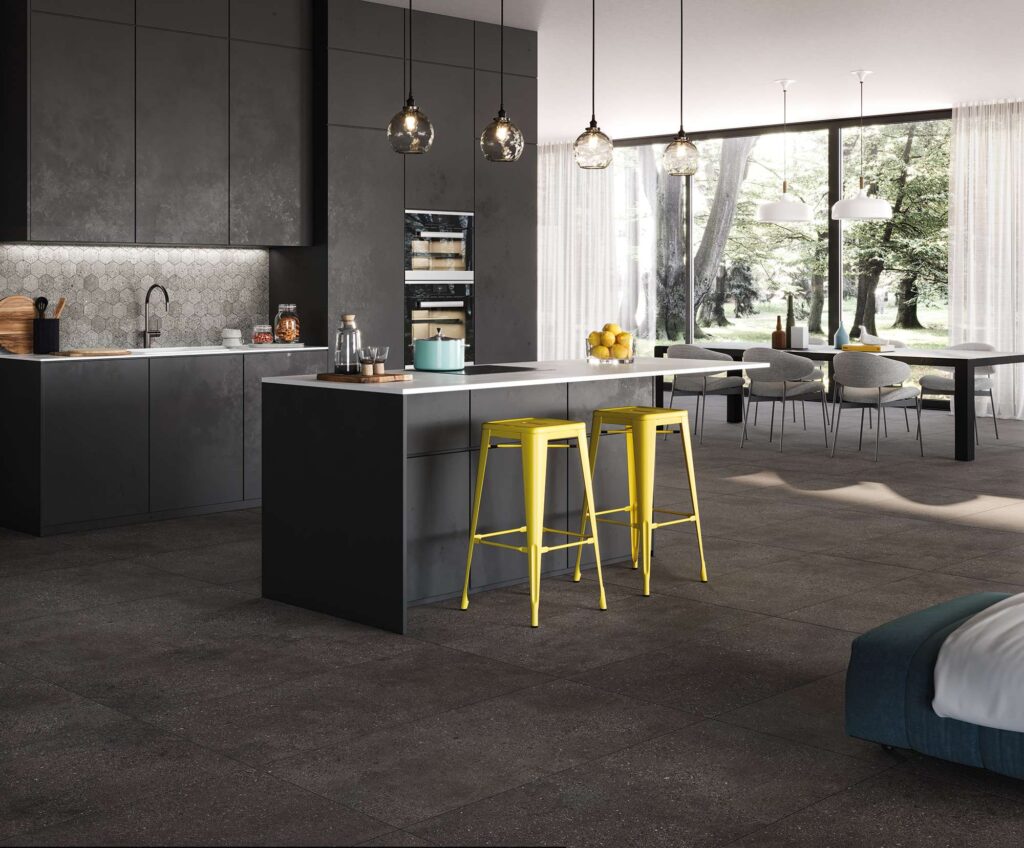 La collezione MON MOKA nel formato 60x120 cm riveste con sobria eleganza il pavimento di un luminoso e moderno open space che unisce cucina e zona living. Le grandi lastre in gres porcellanato color moka scuro, posate in modo regolare e continuo, donano profondità e un raffinato carattere materico all'ambiente. La scena presenta una cucina di design minimalista con isola nera e vivaci sgabelli gialli a contrasto, mentre sullo sfondo ampie vetrate a tutta altezza si affacciano su un parco alberato, inondando lo spazio di luce naturale e valorizzando la texture naturale della pavimentazione.