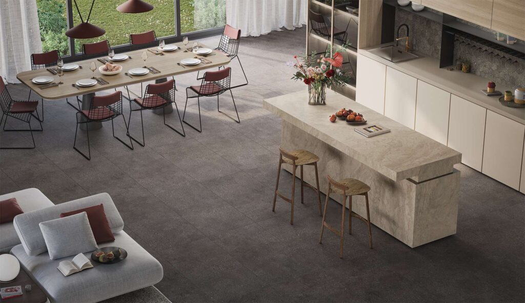 La collezione Mon Moka nel formato 60x120 cm definisce con eleganza il pavimento di questo luminoso e moderno open space. Le piastrelle in gres porcellanato nella calda tonalità marrone scuro moka, posate in modo uniforme, creano un raffinato contrasto visivo con l'isola cucina in pietra chiara e la spaziosa zona pranzo con tavolo in legno. Grandi vetrate affacciate sul giardino e un accogliente angolo soggiorno con divano grigio completano un ambiente dal design contemporaneo, sofisticato e accogliente.