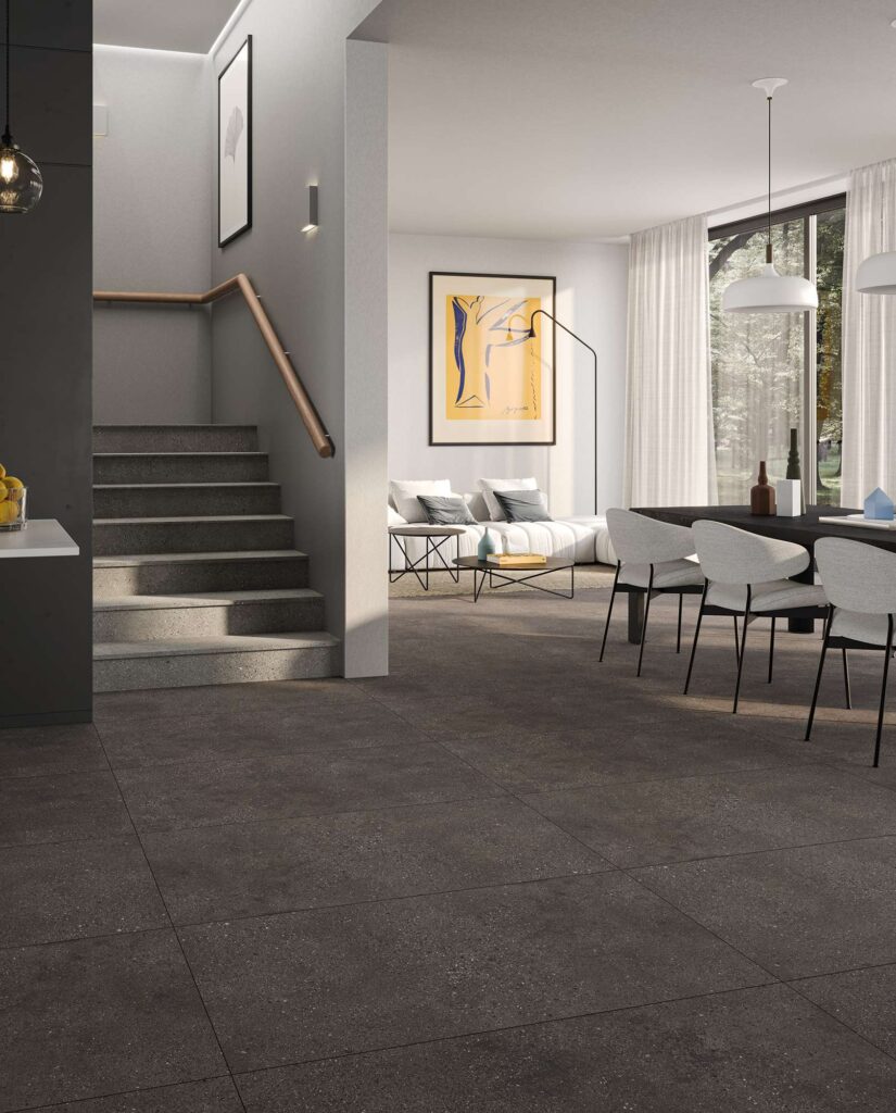 La collezione MON MOKA in formato 60x120 cm veste con eleganza il pavimento di un moderno e luminoso ambiente living open space. Le grandi piastrelle in gres porcellanato nella tonalità moka scuro, caratterizzate da una texture materica e naturale, sono posate a correre, donando continuità visiva allo spazio. Sulla destra, la zona pranzo è arredata con un tavolo scuro, sedie di design bianche e lampadari a sospensione minimalisti; sullo sfondo si scorge un accogliente salotto con divano chiaro e ampie vetrate affacciate sul verde. Sulla sinistra, una scala con gradini in pietra grigia e corrimano in legno aggiunge un tocco di architettura contemporanea e raffinata.