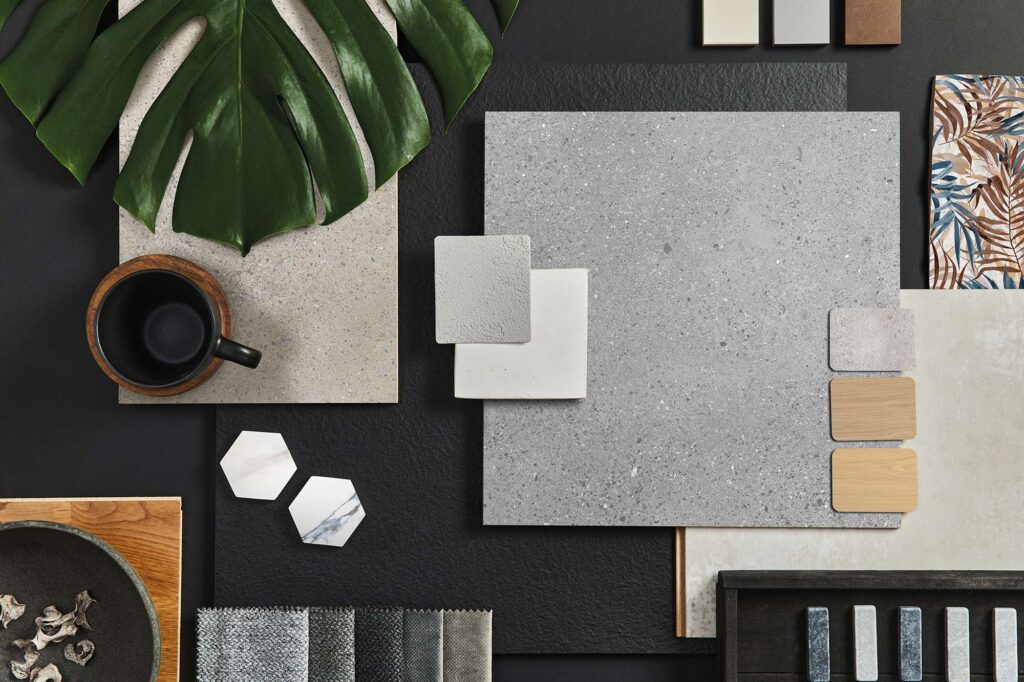 MON MOODBOARD: una raffinata composizione flat lay di ispirazione per il design d'interni, che accosta le piastrelle della collezione in gres porcellanato effetto pietra grigio e beige a superfici materiche scure. La scena mostra una grande piastrella quadrata grigio chiaro, campioni ceramici esagonali effetto marmo e listelli nei toni del legno naturale. Completano l'atmosfera elegante campioni di tessuti grigi, una foglia verde di monstera, una tazza nera su un piattino in legno e una ciotola di design, disposti ordinatamente su uno sfondo antracite.
