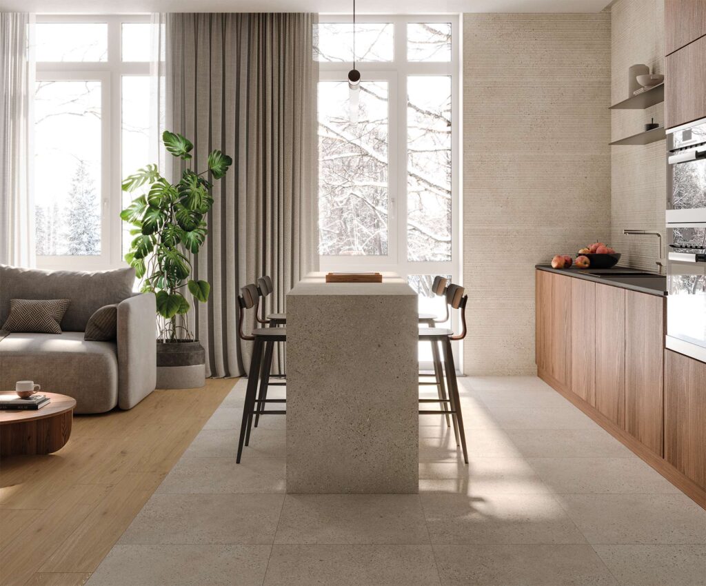 La collezione MON SAND in formato 60x120, nella calda tonalità sabbia, e il decoro a parete DE.LINES valorizzano questo moderno ambiente living e cucina open space di design. Il pavimento in gres porcellanato effetto pietra si coordina armoniosamente con il rivestimento tridimensionale a righe orizzontali sulla parete della cucina. La scena è caratterizzata da una penisola centrale in pietra con sgabelli di design, una zona relax attigua con pavimentazione in legno, divano grigio e una pianta di Monstera, il tutto illuminato dalla luce naturale che filtra dalle ampie finestre affacciate su un paesaggio innevato.