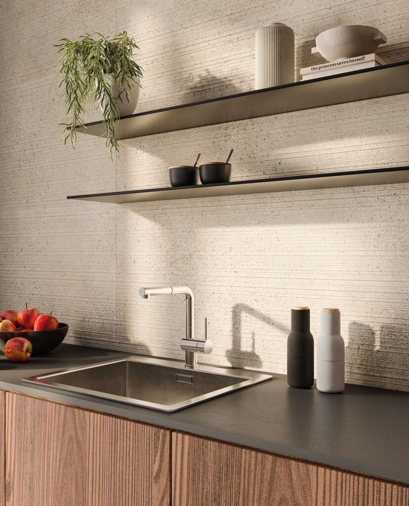 La collezione MON Sand nel formato 60x120 cm, qui proposta nella suggestiva variante decorata Lines, riveste con eleganza la parete backsplash di un moderno ambiente cucina open space. Le grandi lastre ceramiche color sabbia, caratterizzate da una raffinata texture a rilievi orizzontali, creano un'atmosfera calda e materica. Sulla parete sono installate due mensole nere minimaliste con piante ricadenti e oggetti di design, che sovrastano un piano di lavoro scuro con lavello in acciaio inox e miscelatore cromato, completato da basi in legno venato dal gusto contemporaneo.