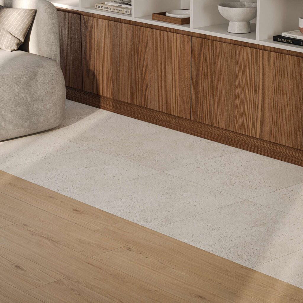 La collezione di piastrelle in gres porcellanato MON SAND nel formato 60X60 si combina con eleganza al gres effetto legno BARRIQUE nella tonalità NAT. HONEY, in questo dettaglio di un raffinato ambiente living. L'immagine evidenzia la posa accostata e la transizione armoniosa tra la superficie chiara e finemente puntinata effetto pietra e le calde doghe effetto legno. Sullo sfondo, un moderno mobile in legno con scaffali a giorno e un divano grigio completano l'atmosfera calda, luminosa e accogliente della stanza.