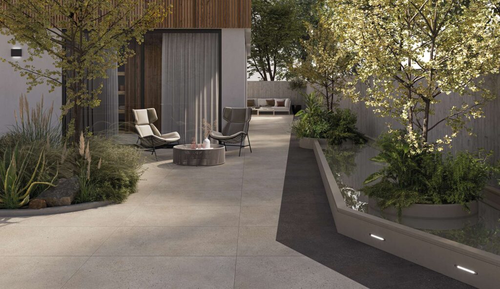 La collezione MON SAND, abbinata alla tonalità Moka nel formato 60x120 cm, definisce la pavimentazione di questo raffinato e moderno ambiente outdoor. L'immagine mostra un elegante patio esterno dove le grandi lastre in gres porcellanato nei toni caldi del sabbia sono posate linearmente, interrotte da una suggestiva fascia scura in contrasto che costeggia uno specchio d'acqua decorativo. Un'area relax con poltrone di design, un tavolino basso, alberi ornamentali e luci integrate crea un'atmosfera lussuosa, accogliente e in perfetta armonia con la natura circostante.
