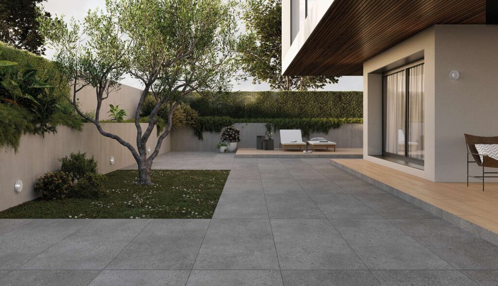 La collezione MON arreda questo raffinato spazio outdoor combinando il gres porcellanato effetto pietra Smoke nel formato 60x120 EXT. con il calore delle doghe effetto legno Barrique Sel. Honey. L'immagine mostra un ampio patio esterno moderno con pavimentazione grigia posata in modo regolare, accostata a camminamenti e pedane rialzate nella tonalità miele. Un ulivo piantato in un piccolo prato, pareti di design con faretti integrati, siepi rigogliose e una zona relax con lettini completano l'atmosfera luminosa e lussuosa di questa residenza contemporanea.