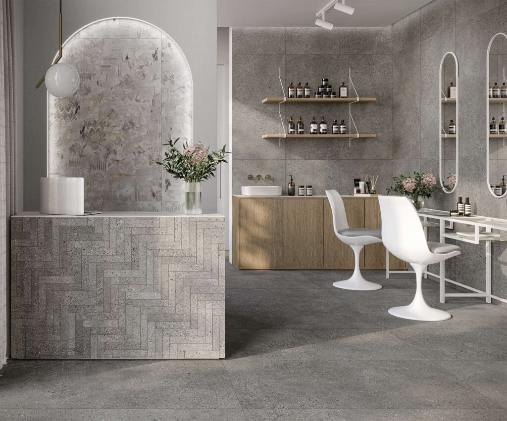 La collezione in gres porcellanato MON SMOKE nel formato 60x120 cm veste con eleganza un moderno salone di bellezza, caratterizzato da superfici grigie effetto pietra naturale. Il pavimento e le pareti sono rivestiti con grandi lastre grigio fumo, mentre il bancone della reception in primo piano presenta una raffinata posa a spina di pesce con la stessa finitura. Sullo sfondo, una nicchia ad arco retroilluminata accoglie il romantico decoro floreale Boheme. L'ambiente, luminoso e sofisticato, è completato da arredi di design minimalisti nei toni del bianco e del legno naturale.
