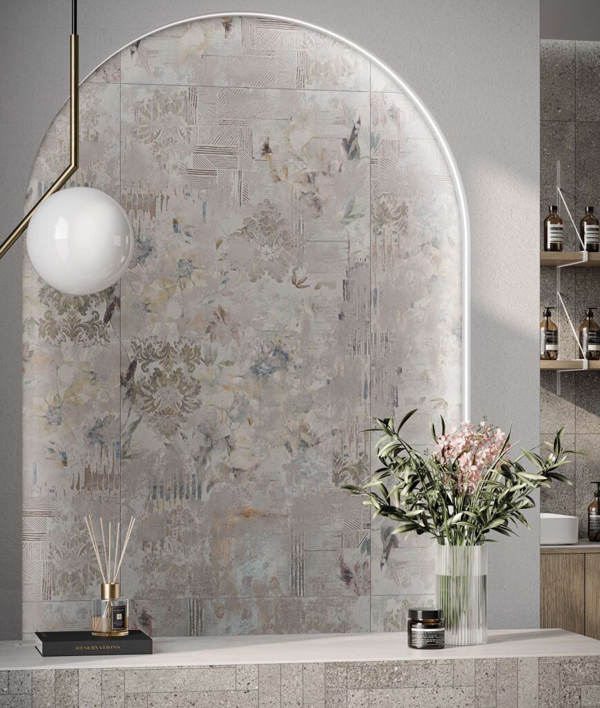 La collezione MON SMOKE, qui abbinata al ricercato decoro BOHEME nel formato 60x120 cm, riveste con eleganza una scenografica nicchia ad arco retroilluminata a LED all'interno di un raffinato salone di bellezza. Le piastrelle in gres porcellanato nei toni del grigio fumo presentano un suggestivo pattern floreale e damascato dall'effetto vintage e vissuto. In primo piano, su un moderno bancone in pietra, trovano spazio un vaso in vetro rigato con rami verdi e fiori rosa, un diffusore di fragranze e una lampada sferica a sospensione dal design minimale, che completano un'atmosfera di lussuoso benessere.