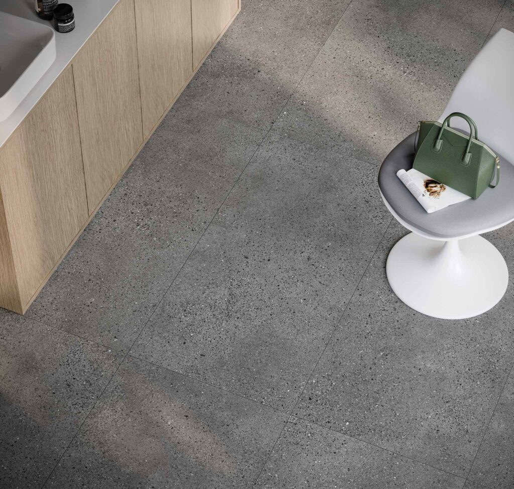 La collezione MON nella tonalità Smoke, nel formato 60x120 cm, si esprime in questa vista dall'alto di un pavimento in gres porcellanato grigio scuro caratterizzato da una raffinata texture materica micropuntinata. La posa regolare delle grandi lastre esalta l'eleganza contemporanea dell'ambiente, perfetto per un salone di bellezza o un'area benessere, qui arredato con un mobile in legno chiaro sulla sinistra e una sedia di design bianca sulla destra.