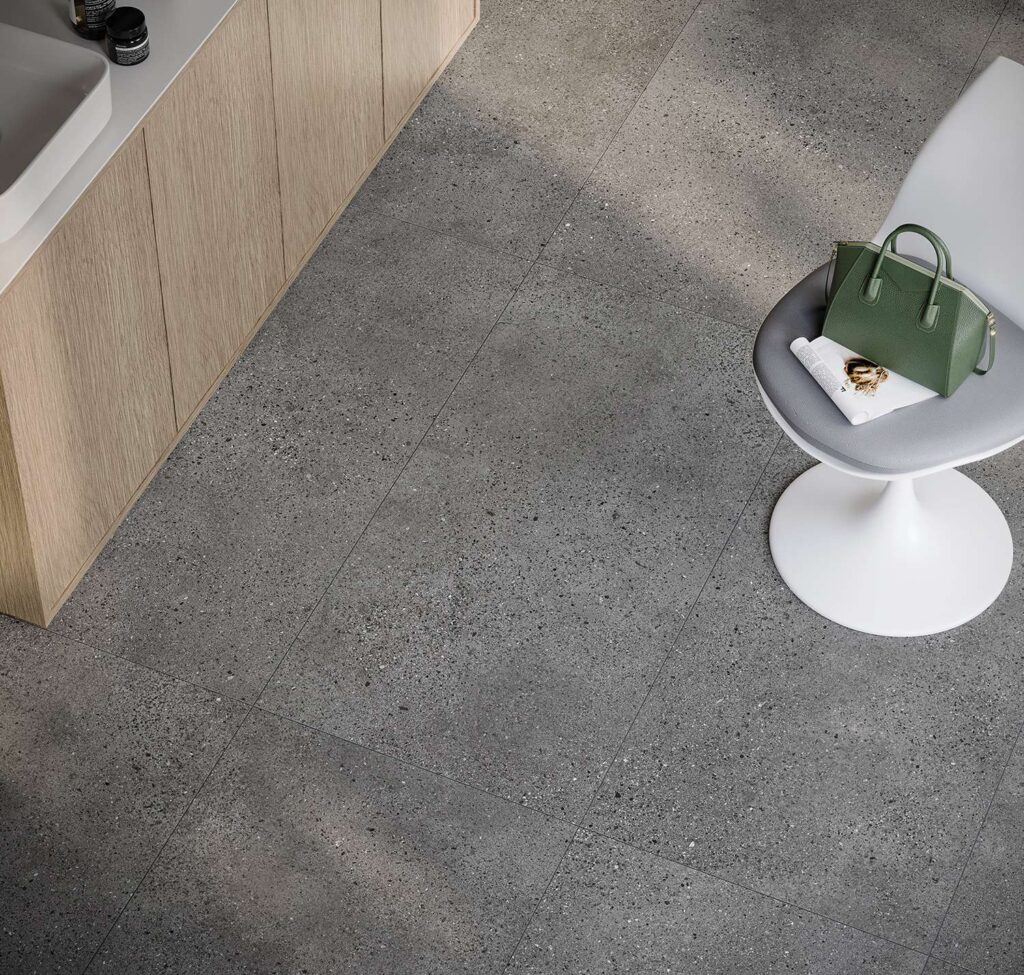 La collezione in gres porcellanato MON SMOKE, qui nel formato 60x120 cm, riveste il pavimento di un ambiente raffinato e contemporaneo con le sue piastrelle color grigio scuro effetto pietra, arricchite da una texture granulare fine e naturale. La posa lineare e geometrica valorizza l'ampiezza dello spazio, in cui si inseriscono arredi minimalisti ed eleganti: a sinistra un mobile sospeso in legno chiaro con lavabo d'appoggio, e a destra una sedia di design bianca con seduta grigia su cui sono adagiate una borsa in pelle verde e una rivista.