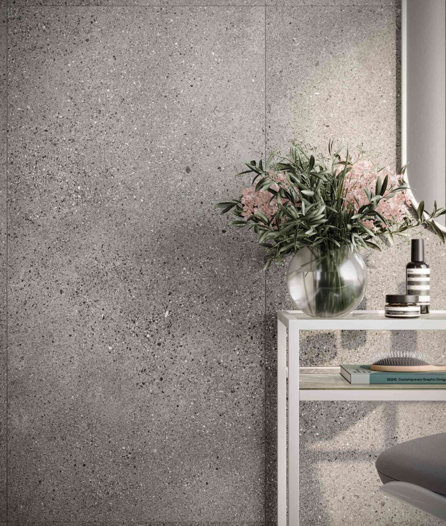 La collezione di piastrelle in gres porcellanato MON SMOKE nel formato 60X120 riveste con eleganza la parete di questo raffinato salone di bellezza, mostrando una texture effetto terrazzo o ceppo di grè nei toni del grigio fumo. La posa verticale a tutta altezza esalta i micro dettagli materici della superficie, accostandosi armoniosamente a una console minimalista bianca su cui poggiano un vaso di vetro con rami d'ulivo e fiori rosa, flaconi di cosmetici e un libro di design, per un'atmosfera di sofisticato benessere.