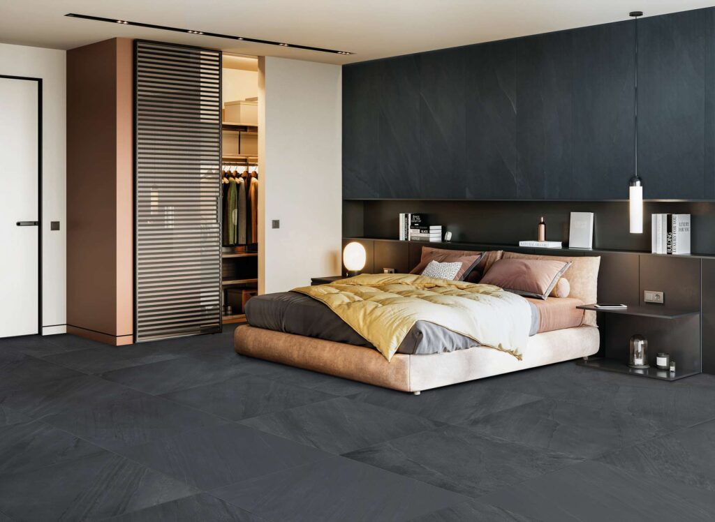 La collezione OCEAN BLACK ABYSS, proposta nei formati 60X120 e 60X60, valorizza una camera da letto moderna dal design ricercato e lussuoso. Il pavimento e il rivestimento della parete retrostante il letto sono realizzati in gres porcellanato nero profondo effetto pietra, regalando una texture materica e un'atmosfera intima. L'ambiente accogliente è completato da un letto matrimoniale con tessuti nei toni dell'oro e del grigio, una cabina armadio visibile sullo sfondo e un'illuminazione calda che esalta le sfumature naturali della ceramica scura.