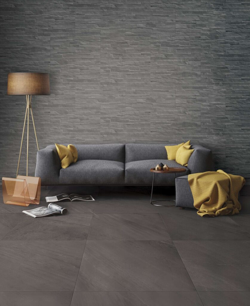 Collezione OCEAN BLACK ABYSS in gres porcellanato effetto pietra scura, qui proposta nel formato 60x60 cm a pavimento e nella variante a muretto come rivestimento per una raffinata parete di fondo. L'elegante ambiente living, caratterizzato da un'atmosfera calda e contemporanea, è arredato con un moderno divano grigio impreziosito da cuscini e dettagli color giallo senape, una lampada da terra a treppiede con luce soffusa e un tavolino da caffè, che esaltano le sfumature intense e materiche della ceramica grigio scuro.