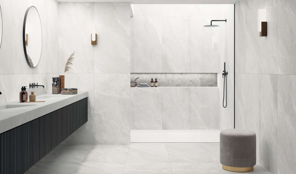 La collezione OCEAN BLACK PEARL veste questo elegante bagno moderno con grandi piastrelle in formato 60x120 cm dall'effetto pietra grigio chiaro e luminoso, posate sia a pavimento che a parete. Nella nicchia della doccia walk-in è inserito un raffinato dettaglio decorativo realizzato con il mosaico in formato 5x5 cm. L'arredamento minimalista, composto da un lungo mobile lavabo sospeso con finitura rigata scura, rubinetteria nera a contrasto e un pouf di design, esalta l'atmosfera luminosa, lussuosa e rilassante dell'ambiente.