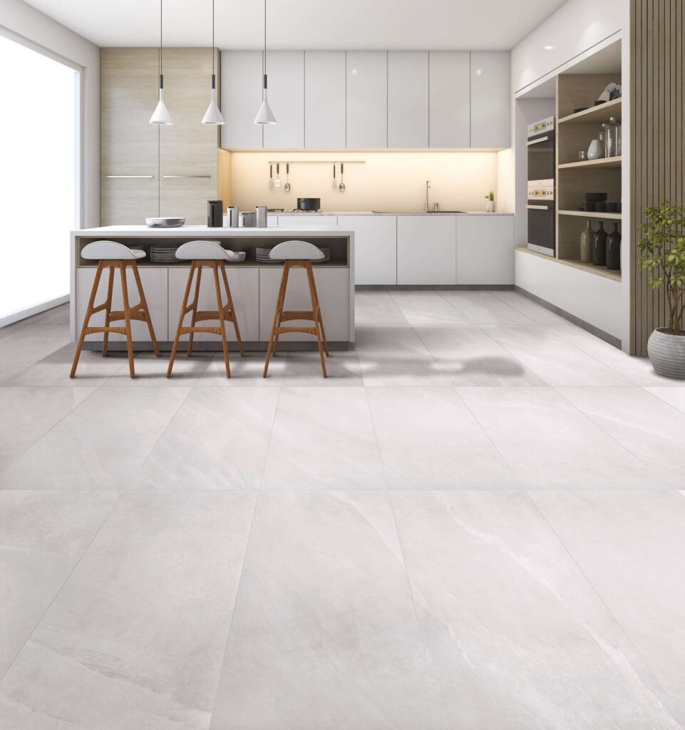 Pavimento in gres porcellanato della collezione Ocean Black Pearl nel formato 60x120, caratterizzato da una tonalità grigio chiaro perlato con delicate venature effetto pietra. Le grandi piastrelle sono posate in modo regolare in un'ampia e luminosa cucina moderna, arredata con pensili bianchi minimalisti, dettagli in legno naturale e un'isola centrale completata da tre sgabelli coordinati.