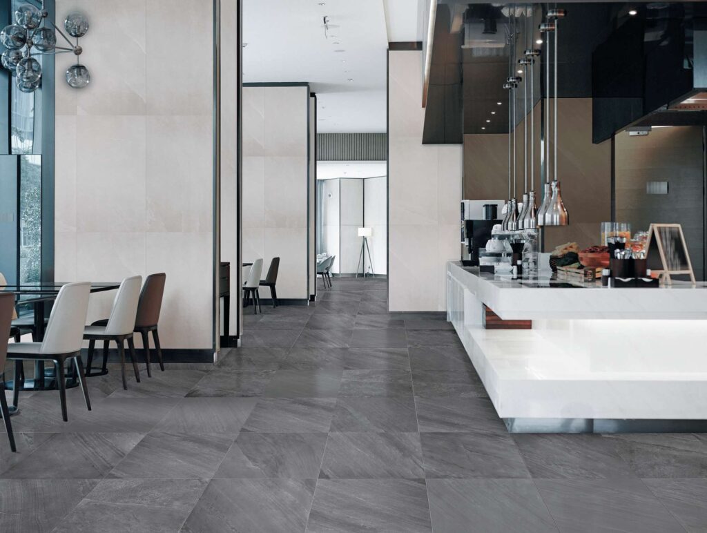Collezione OCEAN BLACK ROCK: pavimento in gres porcellanato effetto pietra scura nel formato 60x60 cm, posato in un elegante e luminoso bar dal design contemporaneo. La vista mostra la texture naturale delle piastrelle grigio scuro che rivestono il pavimento, creando un raffinato contrasto con le pareti chiare, il moderno bancone bianco sulla destra e la zona tavoli con sedie di design sulla sinistra.