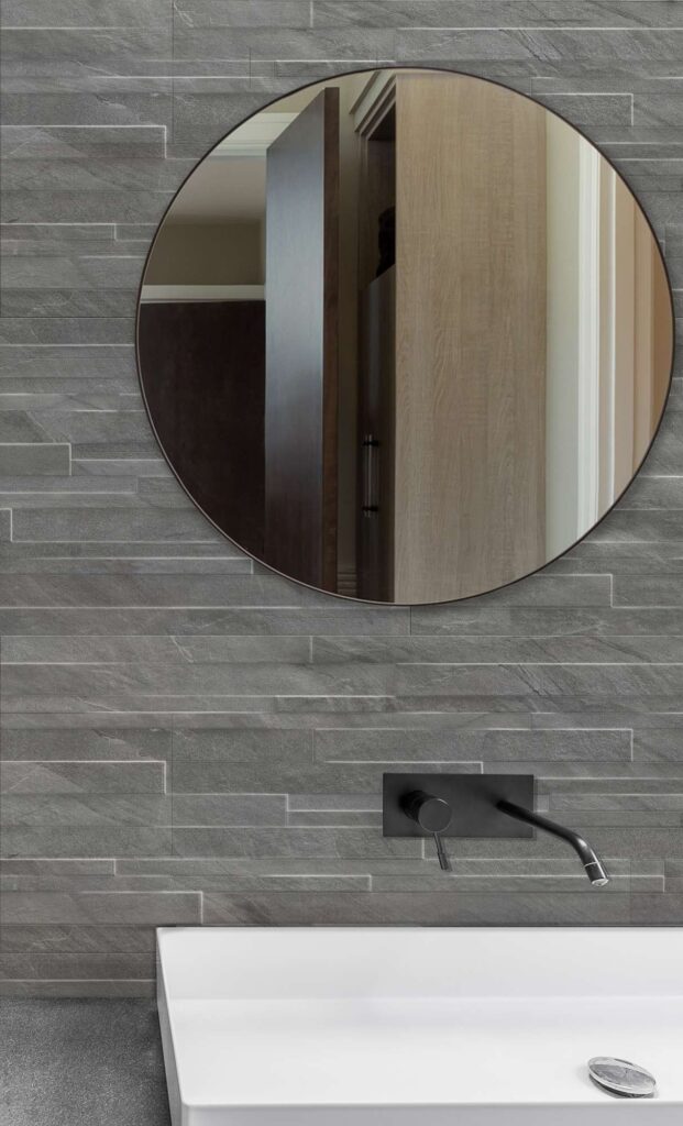 La collezione Ocean Black Rock Muretto nel formato 30x60 cm caratterizza questa parete bagno dal design moderno e raffinato. Il dettaglio ravvicinato mostra la texture tridimensionale del gres porcellanato effetto pietra grigio scuro con posa a muretto, su cui risaltano un elegante specchio circolare, un miscelatore a parete nero opaco e un lavabo bianco minimalista.