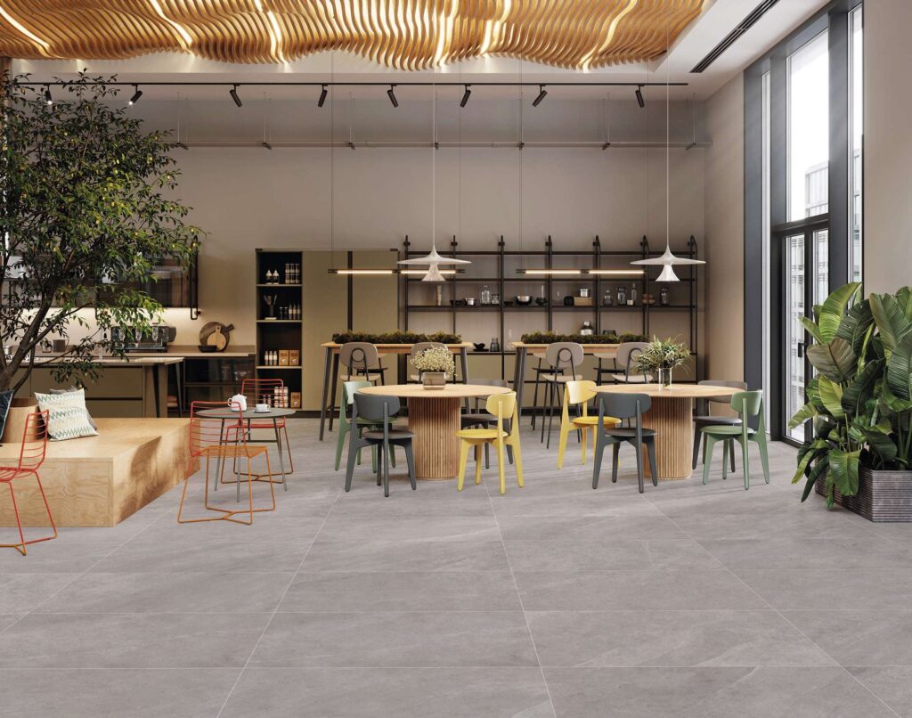 La collezione Ocean Black Wind, nel formato 60x120 cm, riveste con eleganza il pavimento di un moderno e luminoso ristorante. Le piastrelle in gres porcellanato effetto pietra grigia, posate con giunti minimi in modo lineare e continuo, donano un senso di ampiezza e pulizia visiva allo spazio. L'ambiente, dal design contemporaneo e accogliente, è caratterizzato da grandi vetrate, tavoli rotondi in legno completati da sedie colorate, piante ornamentali e un originale soffitto con doghe in legno ondulate che diffonde una luce calda e d'atmosfera.