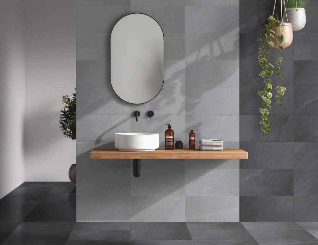 La collezione OCEAN BLACK, proposta nelle varianti colore Wind (grigio chiaro) e Abyss (grigio scuro antracite) nel formato 30x60, valorizza un raffinato ambiente bagno dal design contemporaneo. Le piastrelle in gres porcellanato, posate in senso orizzontale, creano un elegante gioco di contrasti: la tonalità chiara Wind definisce la parete centrale dietro il lavabo a bacinella bianco, posizionato su un piano in legno sospeso, mentre la tonalità scura Abyss riveste la pavimentazione e le pareti laterali. Uno specchio ovale con profilo nero, rubinetteria coordinata minimalista e piante verdi sospese completano questo spazio intimo e luminoso, pervaso da un'atmosfera di sereno benessere.