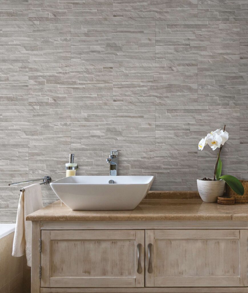 La collezione Ocean Black, qui proposta nella variante Wind Muretto in formato 30x60 cm, decora con eleganza la parete di un bagno raffinato. La posa a muretto delle piastrelle effetto pietra, caratterizzate da delicate sfumature grigie e beige, crea uno sfondo materico e naturale dietro a un moderno lavabo a bacinella bianco, posizionato su un mobile in legno chiaro con top in pietra. Un'orchidea bianca e dettagli curati completano l'atmosfera rilassante e di design dell'ambiente.