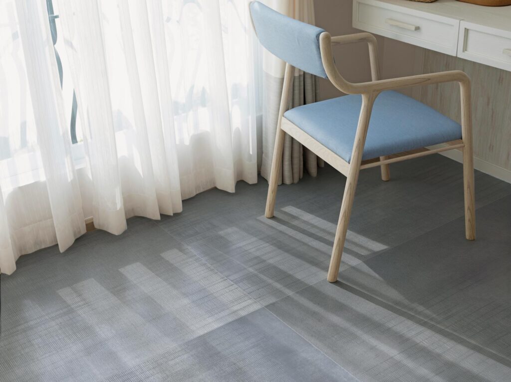 Collezione LIVING: pavimento in gres porcellanato effetto tessuto nella sofisticata tonalità Denim, formato 60x120 cm. L'immagine mostra un luminoso angolo di un ambiente residenziale, dove la luce del sole filtra attraverso tende bianche e leggere, proiettando eleganti ombre geometriche sulla superficie materica delle piastrelle. Una sedia di design in legno chiaro con seduta azzurra è accostata a uno scrittoio bianco, creando un'atmosfera serena, moderna e raffinata che valorizza la trama e la posa del pavimento.