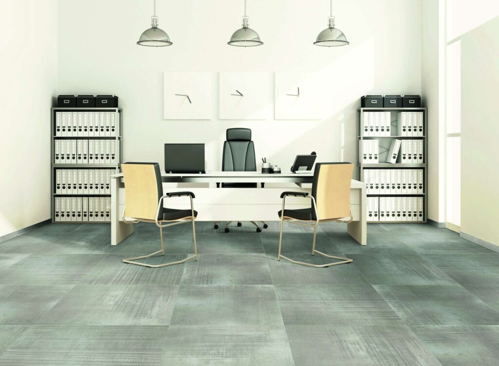 La collezione di piastrelle RUG SMOKE, nella tonalità grigio fumo con un raffinato effetto tessuto, riveste a pavimento un moderno e luminoso ufficio professionale con grandi lastre di formato quadrato. L'immagine mostra un elegante studio direzionale caratterizzato da una scrivania bianca centrale con sedie di design, scaffalature metalliche ordinate con faldoni bianchi e tre lampade a sospensione dallo stile industriale. La texture ceramica del pavimento evoca la trama di un tappeto contemporaneo, conferendo calore e un'atmosfera di alto livello estetico all'ambiente di lavoro.