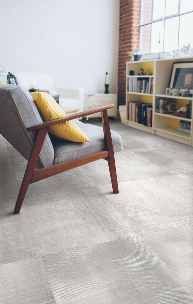 La collezione RUG SMOKE presenta un pavimento in gres porcellanato effetto tessuto nella tonalità grigio Smoke, posato in un moderno e luminoso salotto. L'inquadratura ravvicinata mette in risalto la texture materica e il formato quadrato delle piastrelle, mentre l'ambiente è impreziosito da una poltrona di design in legno e tessuto grigio con un cuscino giallo a contrasto, completata sullo sfondo da una libreria chiara e una parete con mattoni a vista.