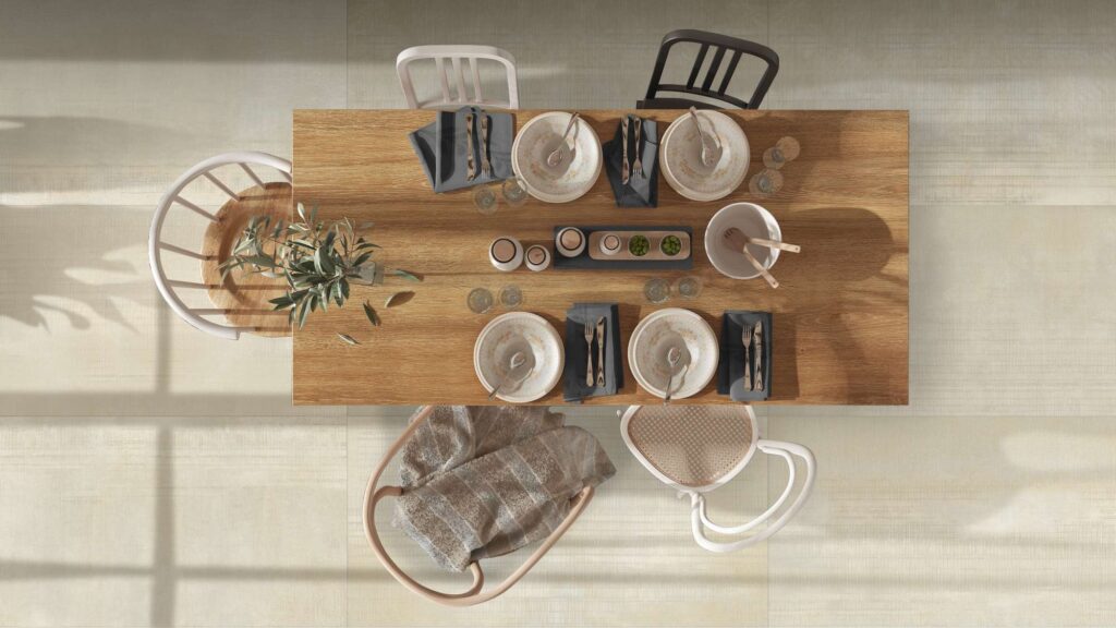 Collezione Organic Rug nella tonalità Sand: pavimento in gres porcellanato dall'effetto naturale e materico, posato in grandi lastre rettangolari e ritratto in una raffinata vista dall'alto. Al centro della scena, un tavolo in legno massello è elegantemente apparecchiato per quattro ospiti con stoviglie coordinate, posate e calici, circondato da sedie di design in stile rustico-contemporaneo. La luce naturale che filtra lateralmente proietta ombre morbide sul pavimento, esaltando la texture calda e l'atmosfera accogliente e sofisticata dell'ambiente pranzo.