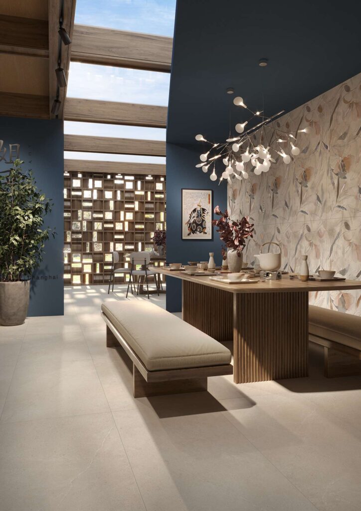 La collezione PALLADIO si esprime in questo elegante ristorante attraverso la posa a pavimento di piastrelle in gres porcellanato beige nel formato 60x120 cm. L'ambiente, dall'atmosfera raffinata e d'ispirazione contemporanea, è caratterizzato da un grande tavolo da pranzo in legno con dettagli cannettati e panche coordinate con seduta imbottita beige. Sulla destra, la parete è arricchita da un rivestimento decorativo con delicati motivi floreali e botanici, accostata a pareti blu notte. Un moderno lampadario scultoreo a rami luminosi diffonde una luce calda sul tavolo apparecchiato, mentre lo sfondo rivela una suggestiva parete divisoria geometrica in legno e un soffitto con lucernario che inonda lo spazio di luce naturale.