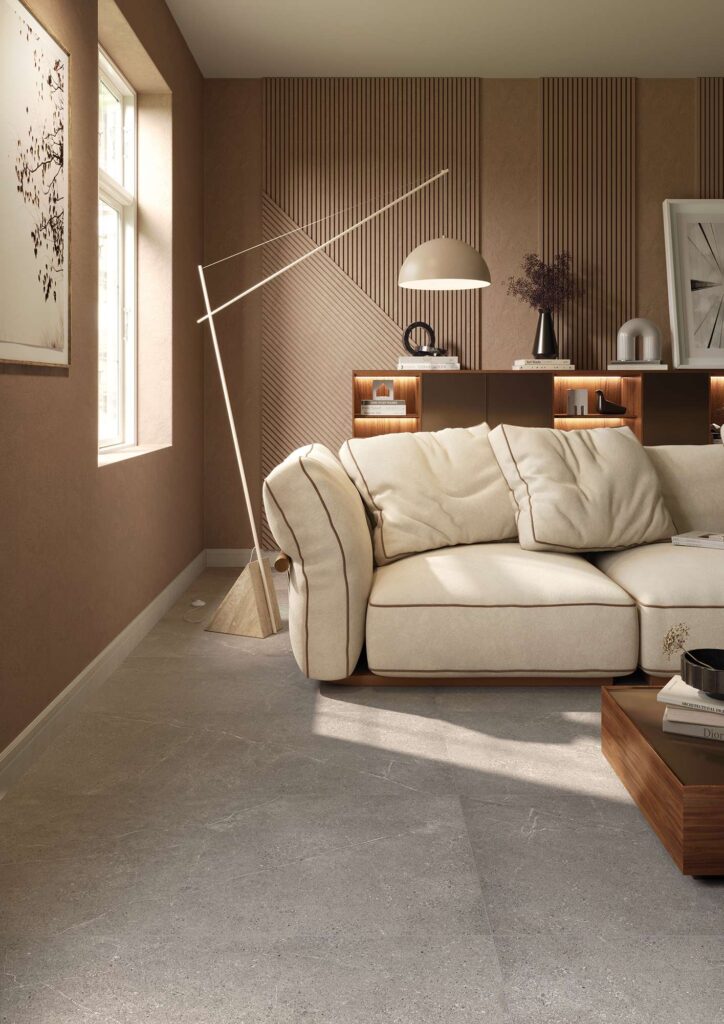 La collezione PALLADIO veste un elegante living moderno con le piastrelle in gres porcellanato color Cenere nel formato 60x120. La pavimentazione effetto pietra grigia, caratterizzata da venature sottili e naturali, si abbina armoniosamente a un arredamento sofisticato nei toni caldi del terracotta e del marrone. L'ambiente accogliente è completato da un grande divano in tessuto crema con profili a contrasto, una parete decorata con boiserie in legno a listelli, una lampada di design e una luce naturale che esalta la superficie materica del pavimento.