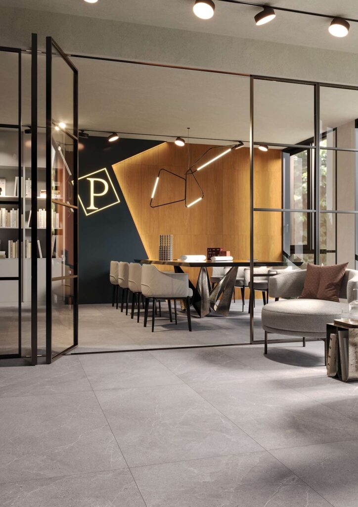 Collezione PALLADIO in gres porcellanato color Grafite, nel formato 60x120, posata a pavimento in un elegante ufficio di rappresentanza. L'immagine mostra le grandi lastre grigie con delicate venature effetto pietra che donano continuità visiva agli spazi, separati da una raffinata vetrata con profili scuri. Oltre la porta a vetri, si apre una sala riunioni moderna e accogliente, arredata con un grande tavolo di design, poltroncine chiare, una parete con boiserie in legno caldo e una sofisticata illuminazione sospesa.